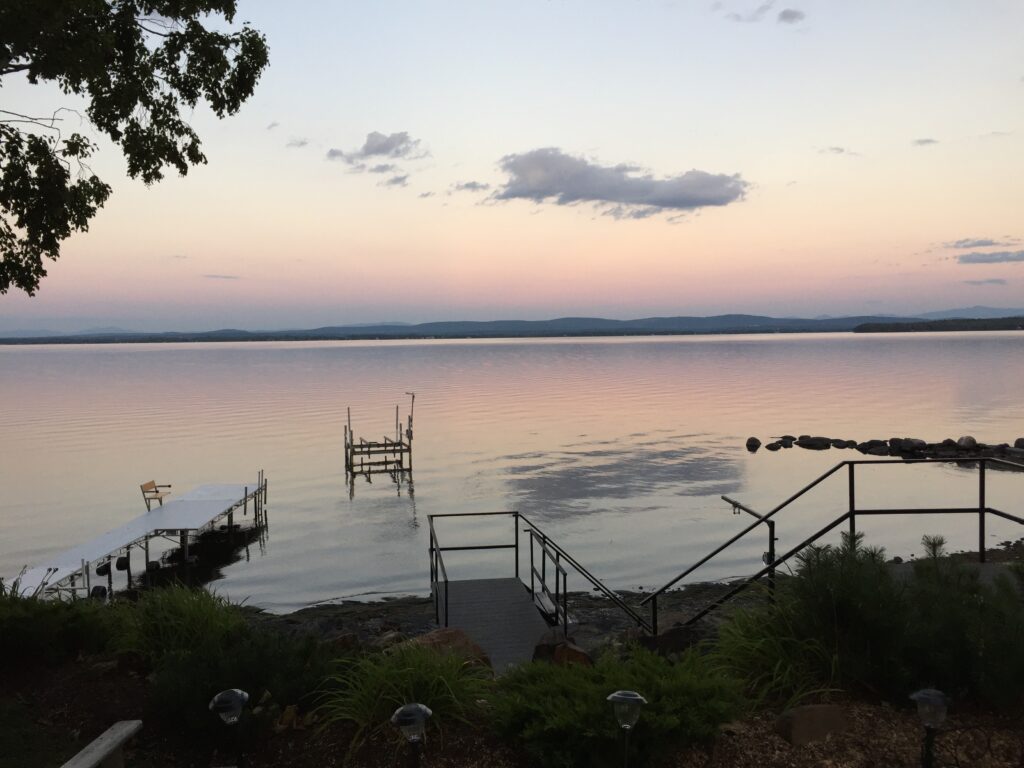 A picture of Lake Champlain as seen from the shore with a dock and boat launch in the water, a rocky point and the Green Mountains in the background.