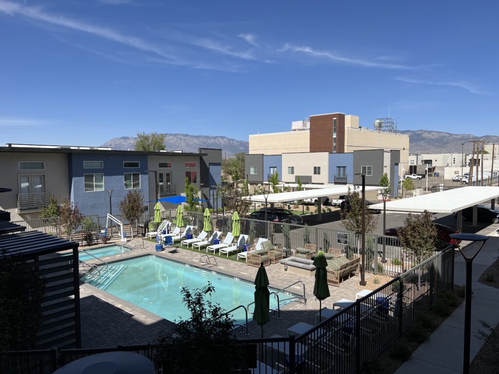 A picture of a swimming pool, hot tub, and lounge chairs in the courtyard of a apartment building as seen from a second floor balcony. There are mountains in the background.